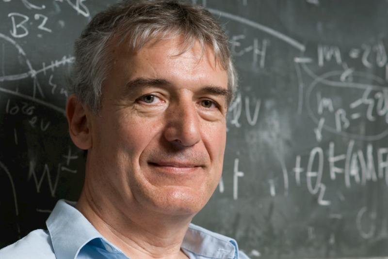 A head shot of professor Sikivie standing in front of a chalkboard with physics equations written on it.