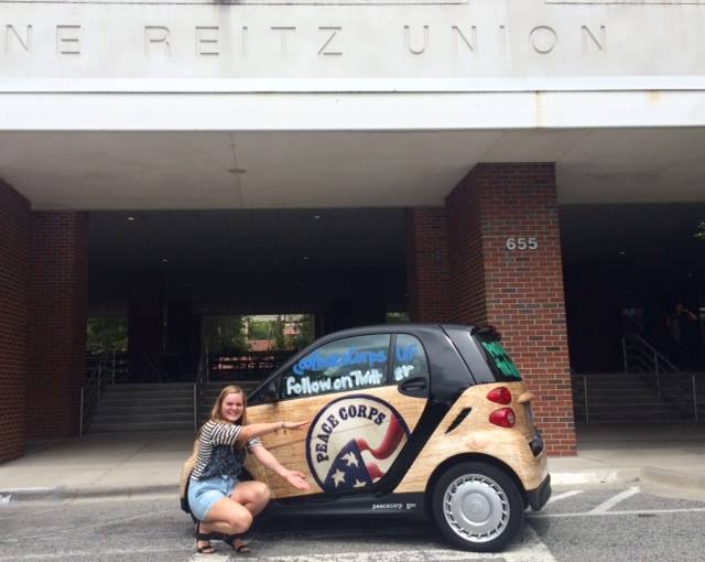 A student poses next to a car with the Peace Corps logo on it.