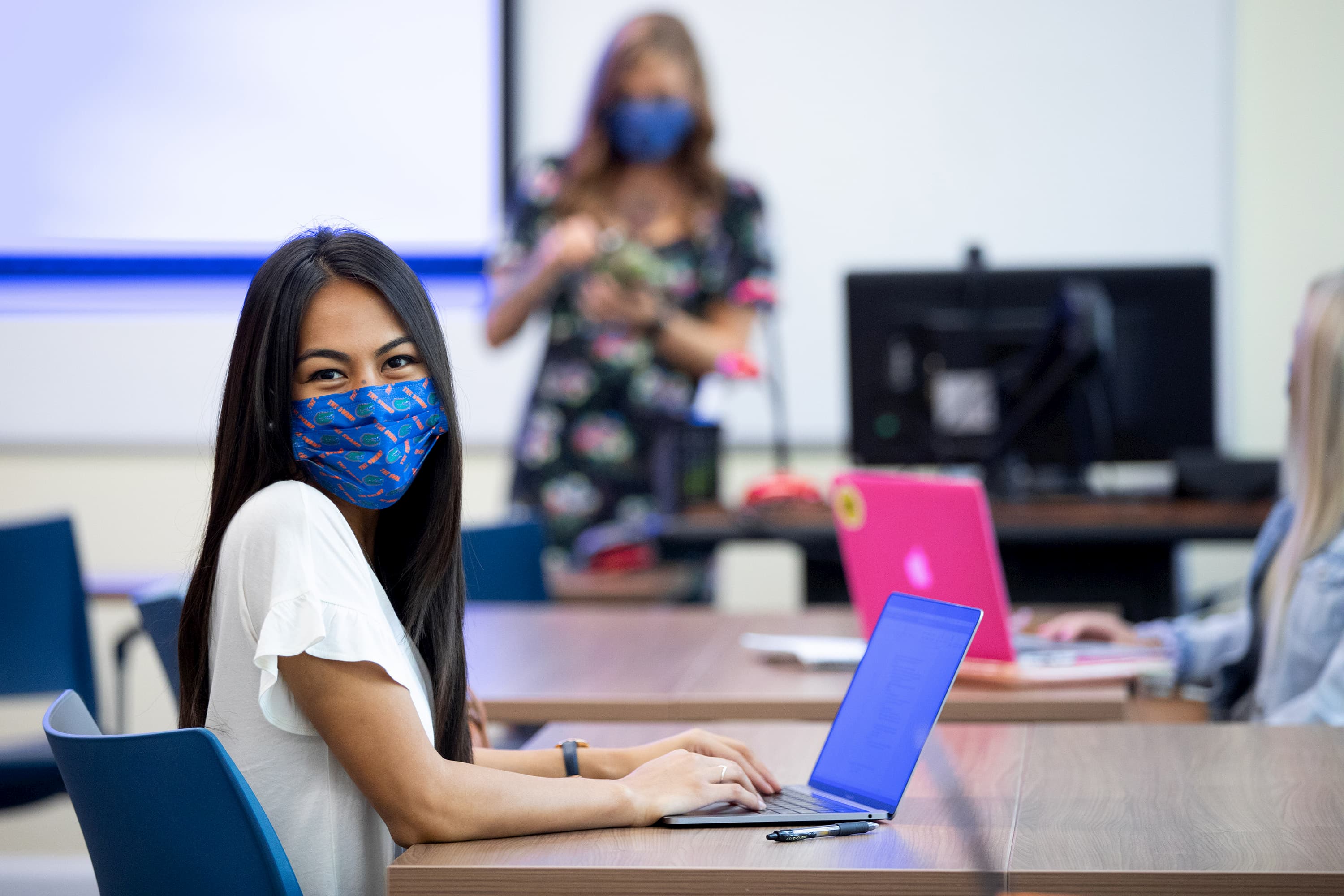 A student sits in a lecture hall wearing a mask while typing on a laptop.