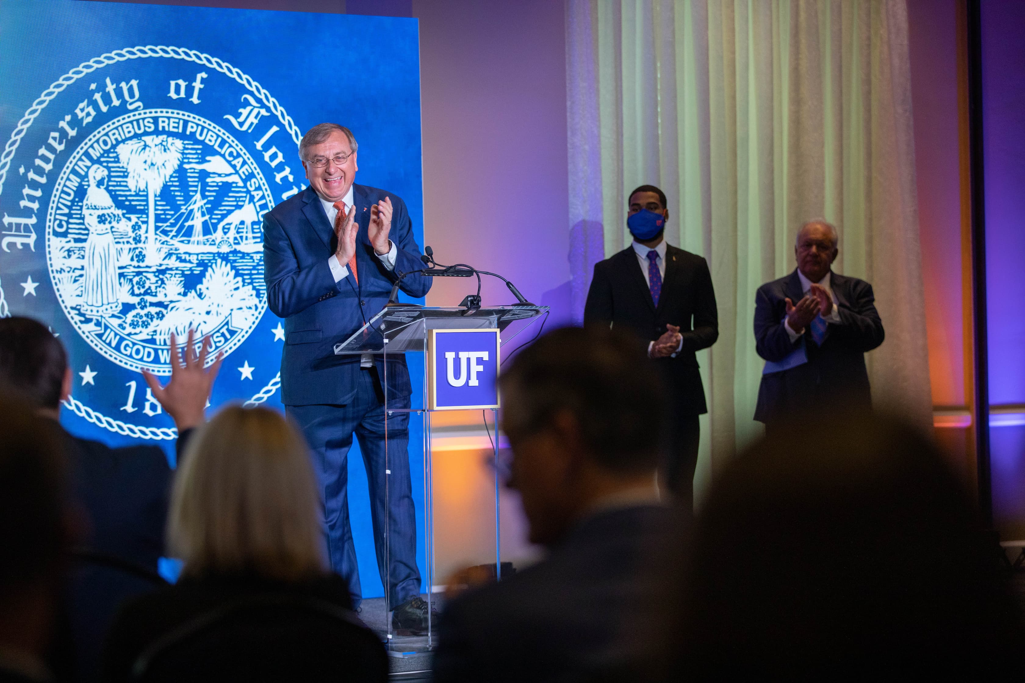 President Fuchs stands in front of a wooden lectern with the blue-and-white UF logo on the front of it. He is clapping and flanked by two members of the UF Board of Trustees.