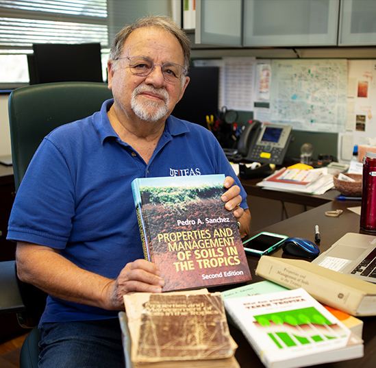 Pedro Sanchez poses for a photo while sitting behind a desk holding one of the books he authored.