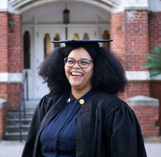 Yesenia Arroyo laughs while wearing a graduation cap and gown on UF's campus. 