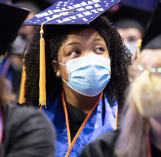 A student wears a graduation cap and gown while sitting in a crowd of other graduates at commencement. She is wearing a surgical mask. 