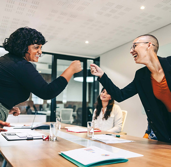 Two women fist bump over an office table in a conference room