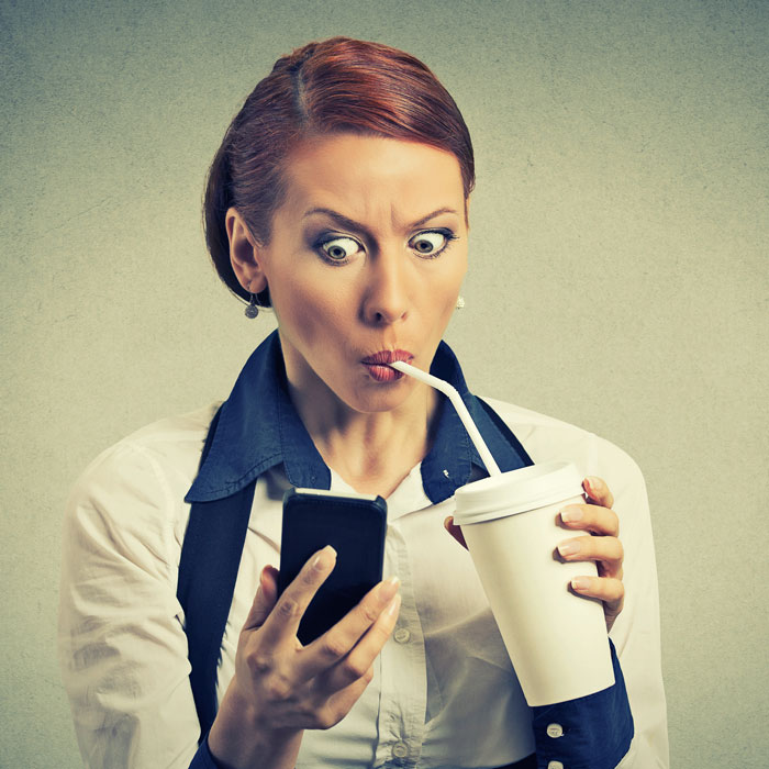 A stock image of a woman sipping on a drink while staring at a her phone with a stunned look.