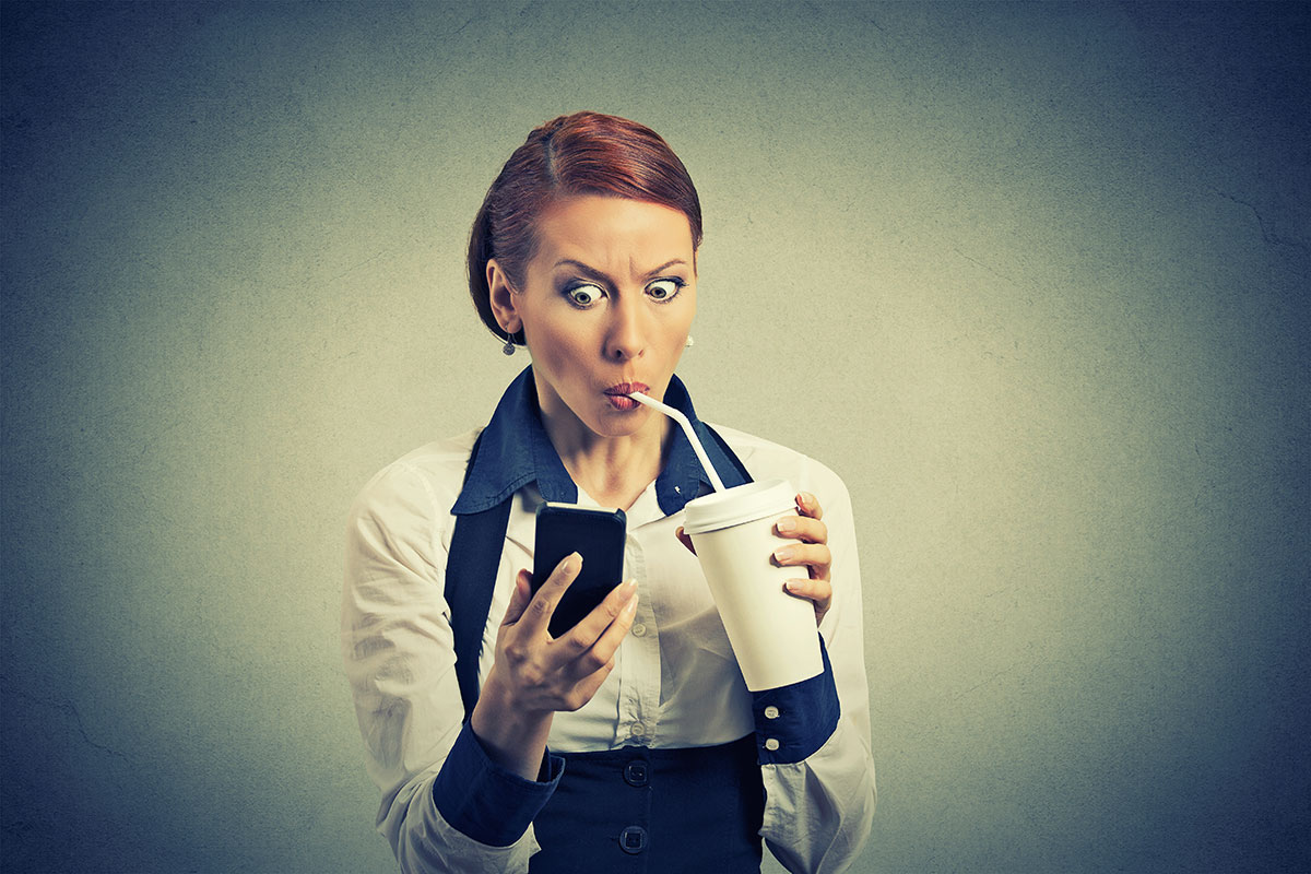 A stock image of a woman sipping on a drink while staring at a her phone with a stunned look.