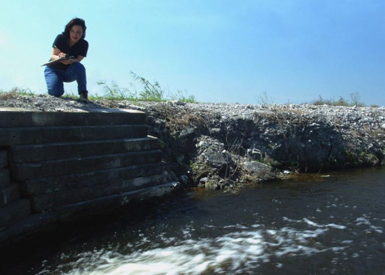 A woman crouches near the bank of a body of water taking notes on a pad
