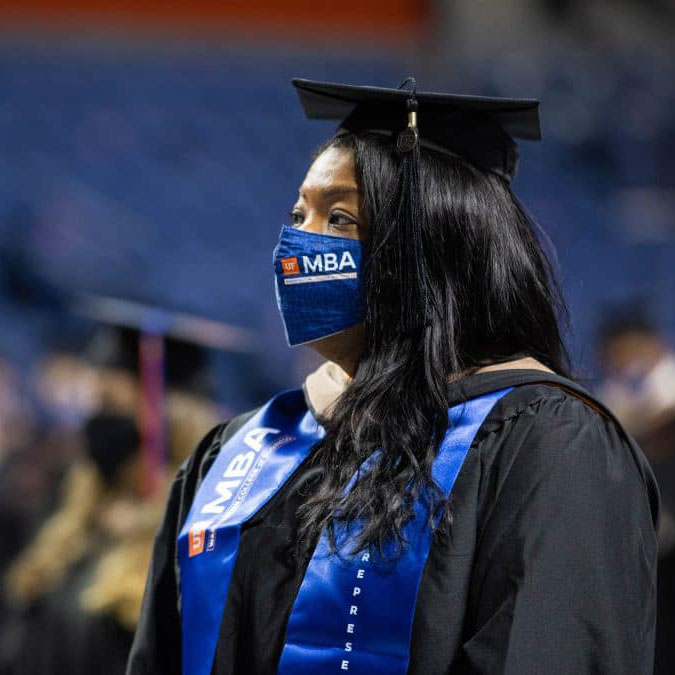 A woman dressed in a university cap & gown while wearing a face mask.