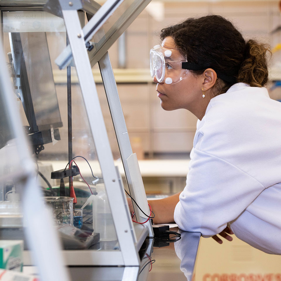 A woman wearing lab goggles and coat looks at laboratory equipment