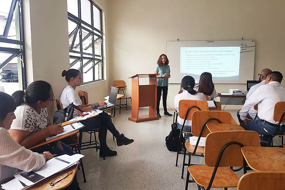 Female teacher stands before several students sitting in their desks in a classroom. 