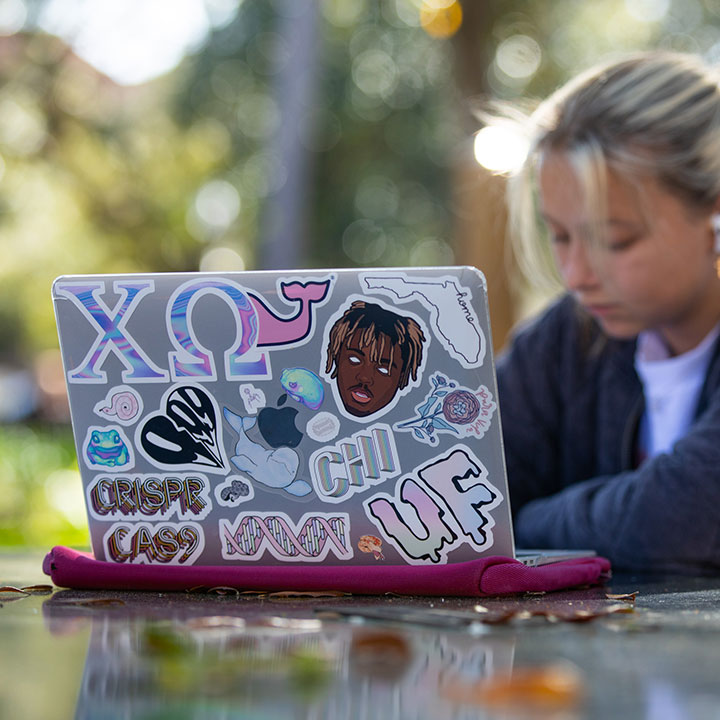 Focal point: a laptop with over a dozen stickers. A student works in the background.