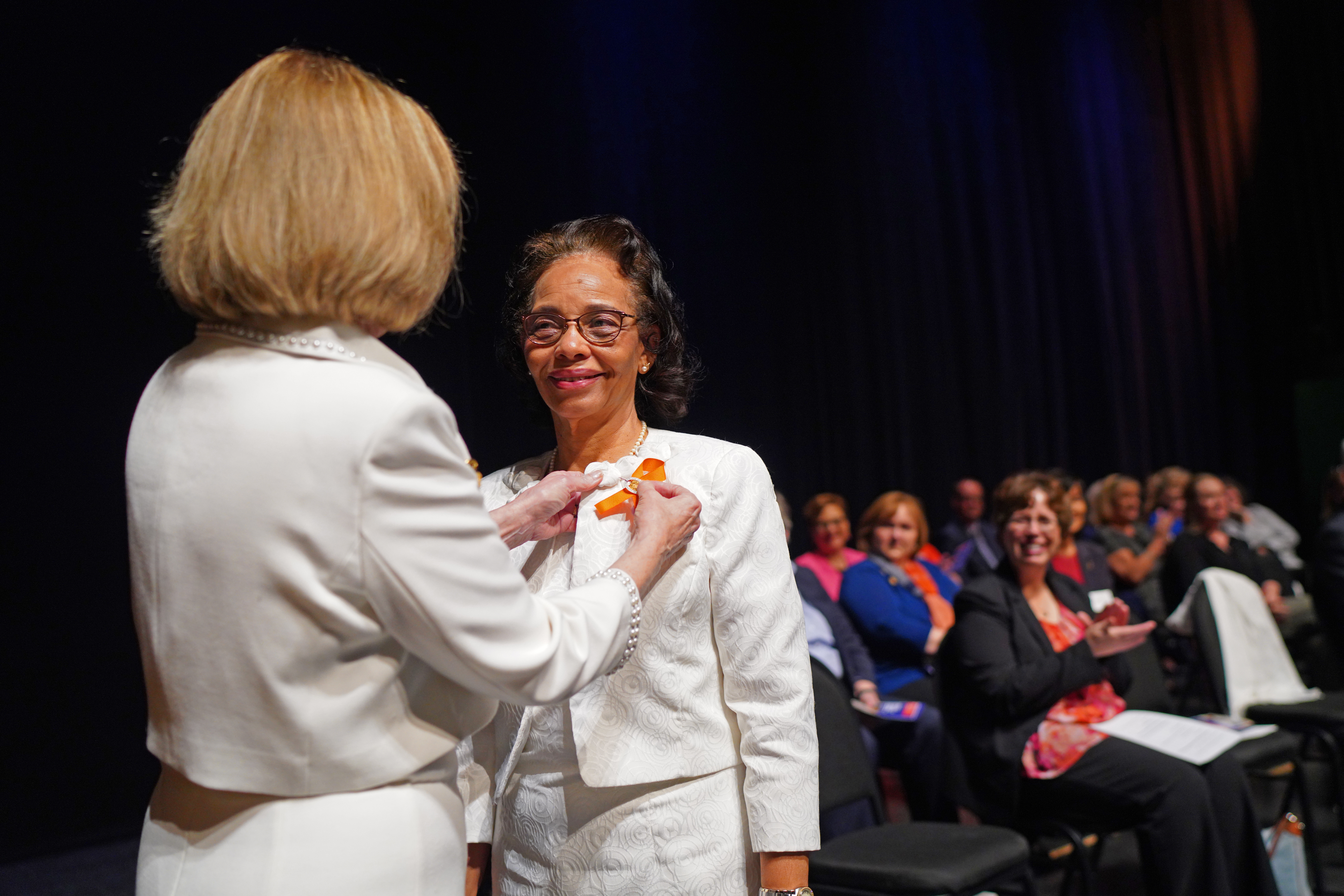 A woman is pinning another woman's shirt at a ceremony on stage. 