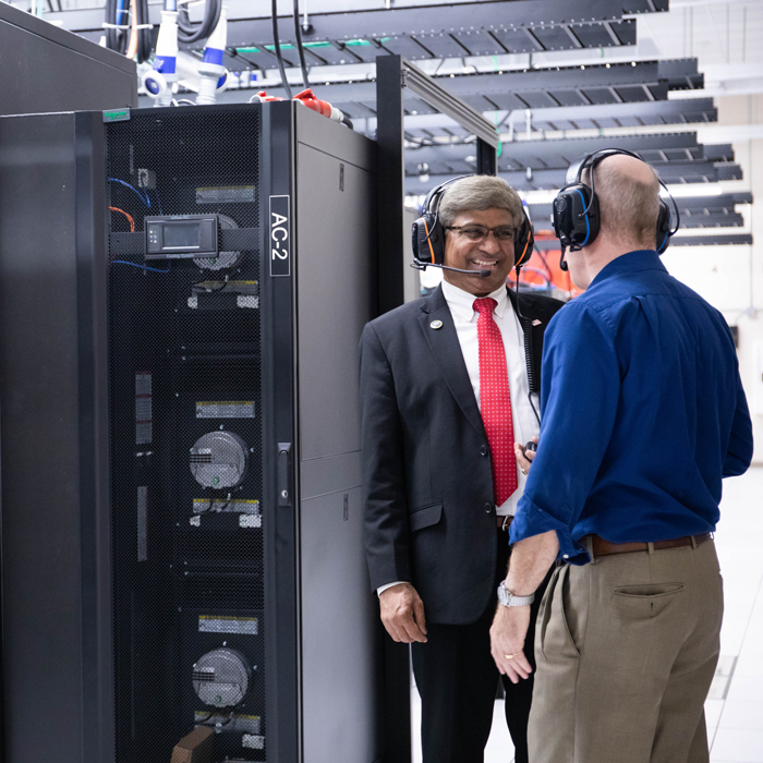 Two men in headphones talk in front of a supercomputer.