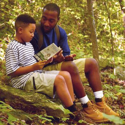 A father and son sit on a trail reading a guide. 