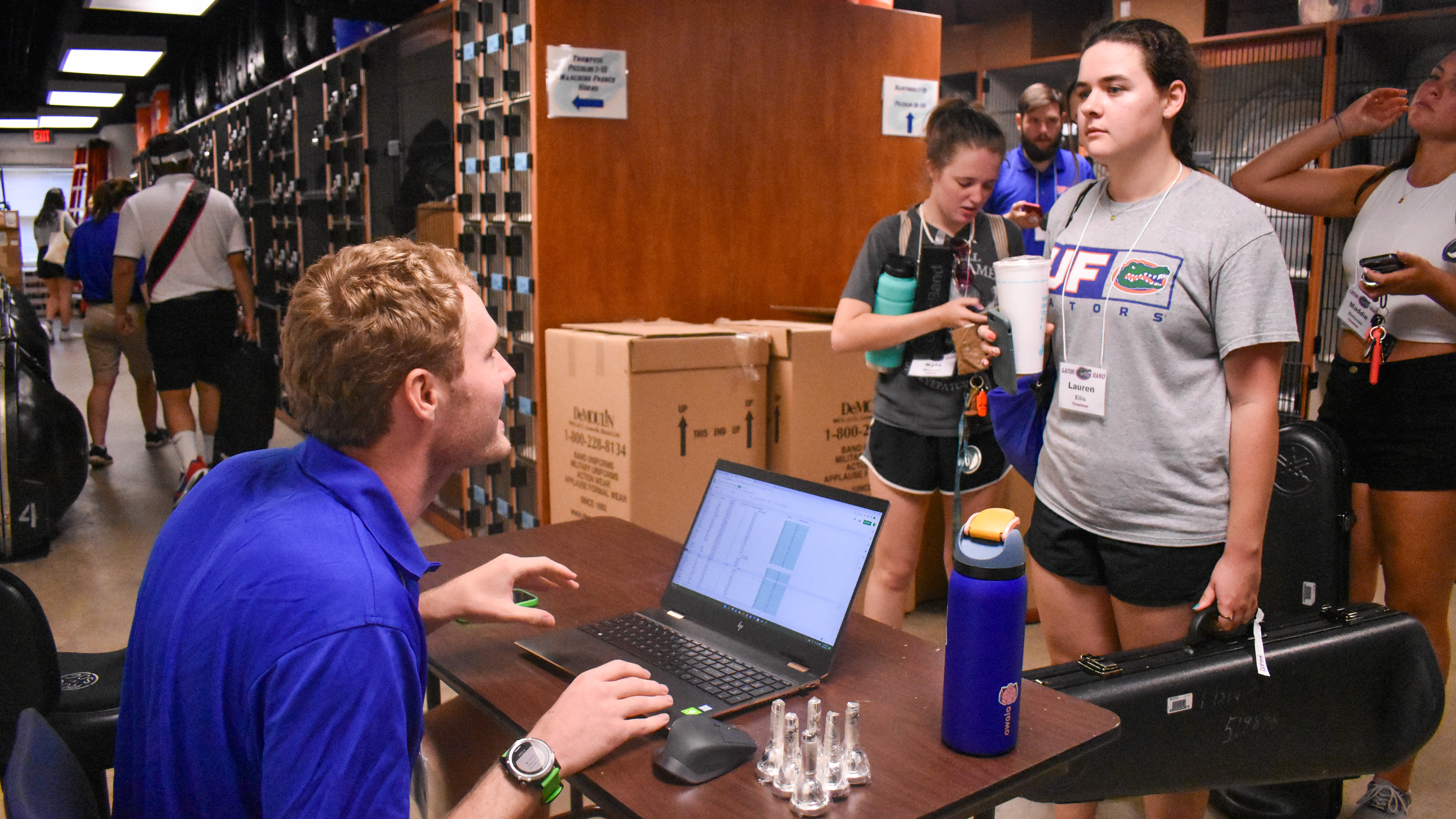 A student at a table types on a computer while talking to someone standing up.