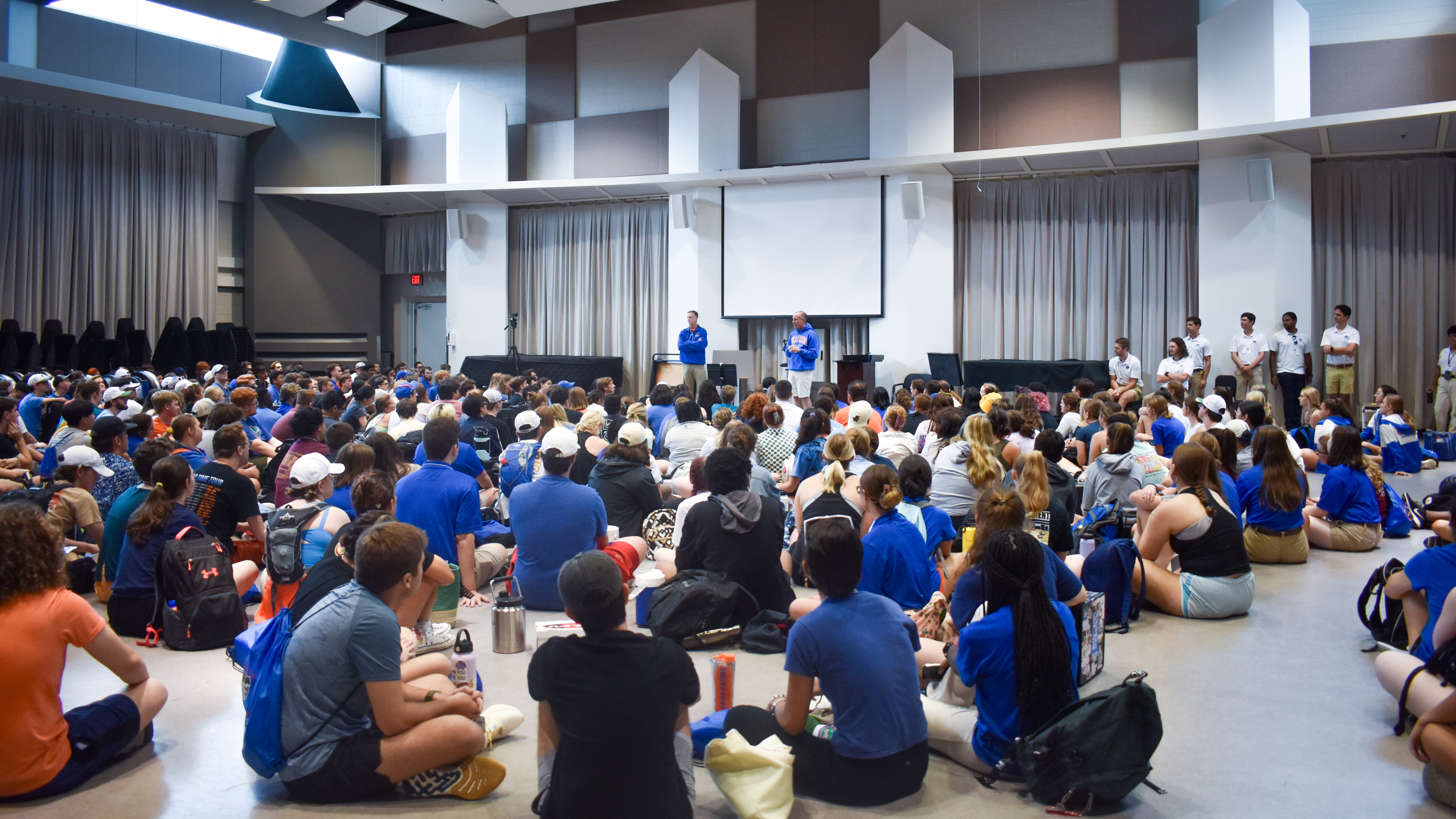 A large group of students sit on the floor inside listening to two professors speak.