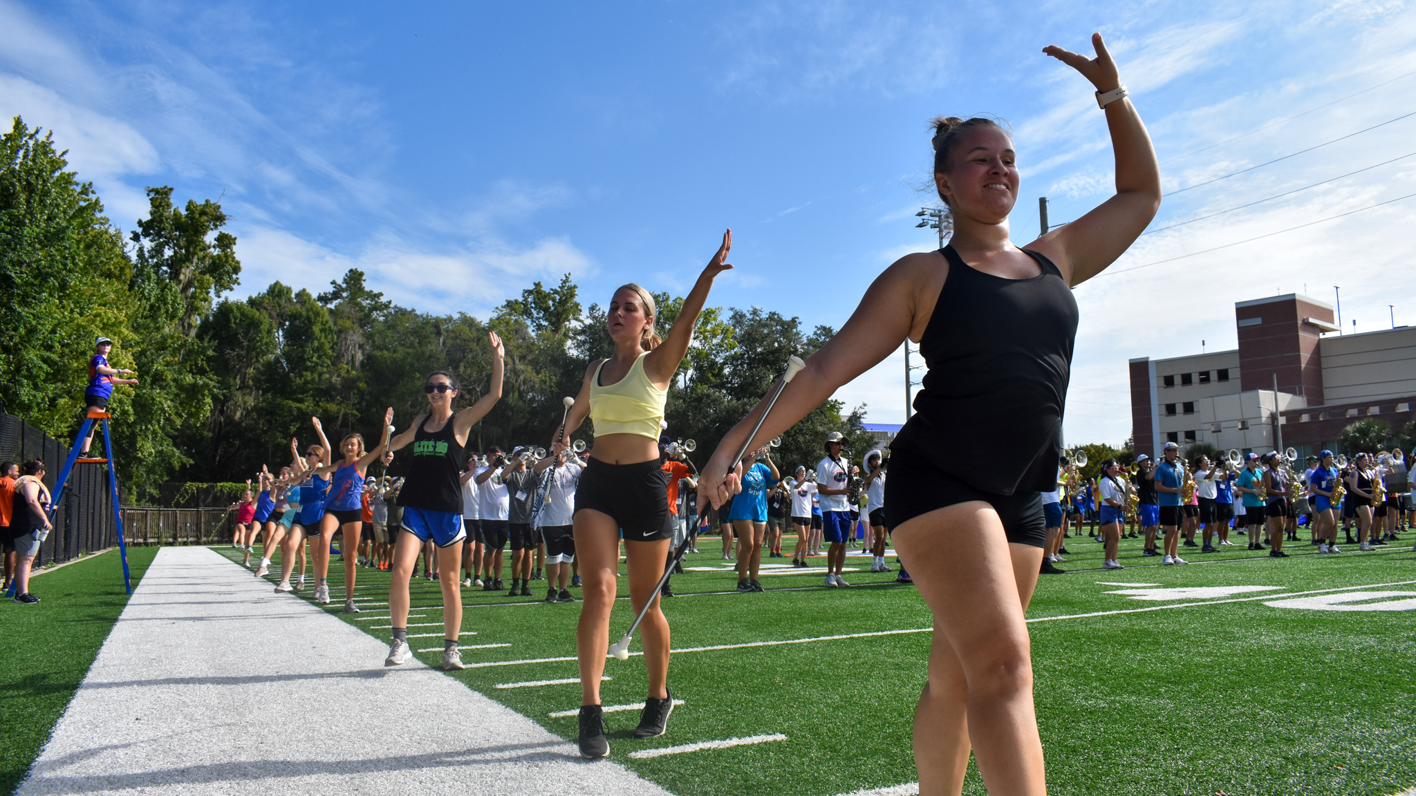 A line of baton twirlers with a marching band in the background.