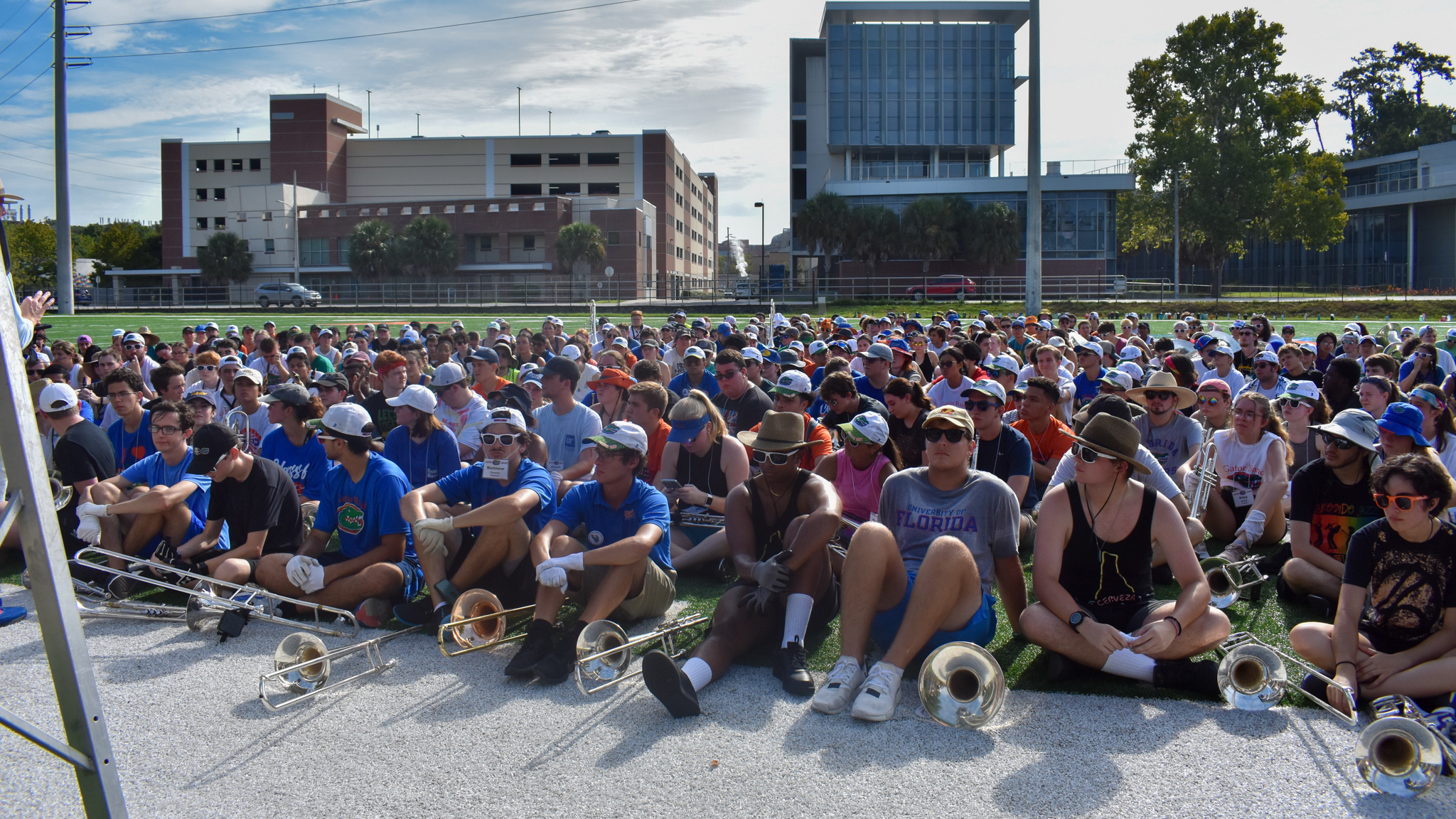 A marching band sits on a football field and listens to a professor.