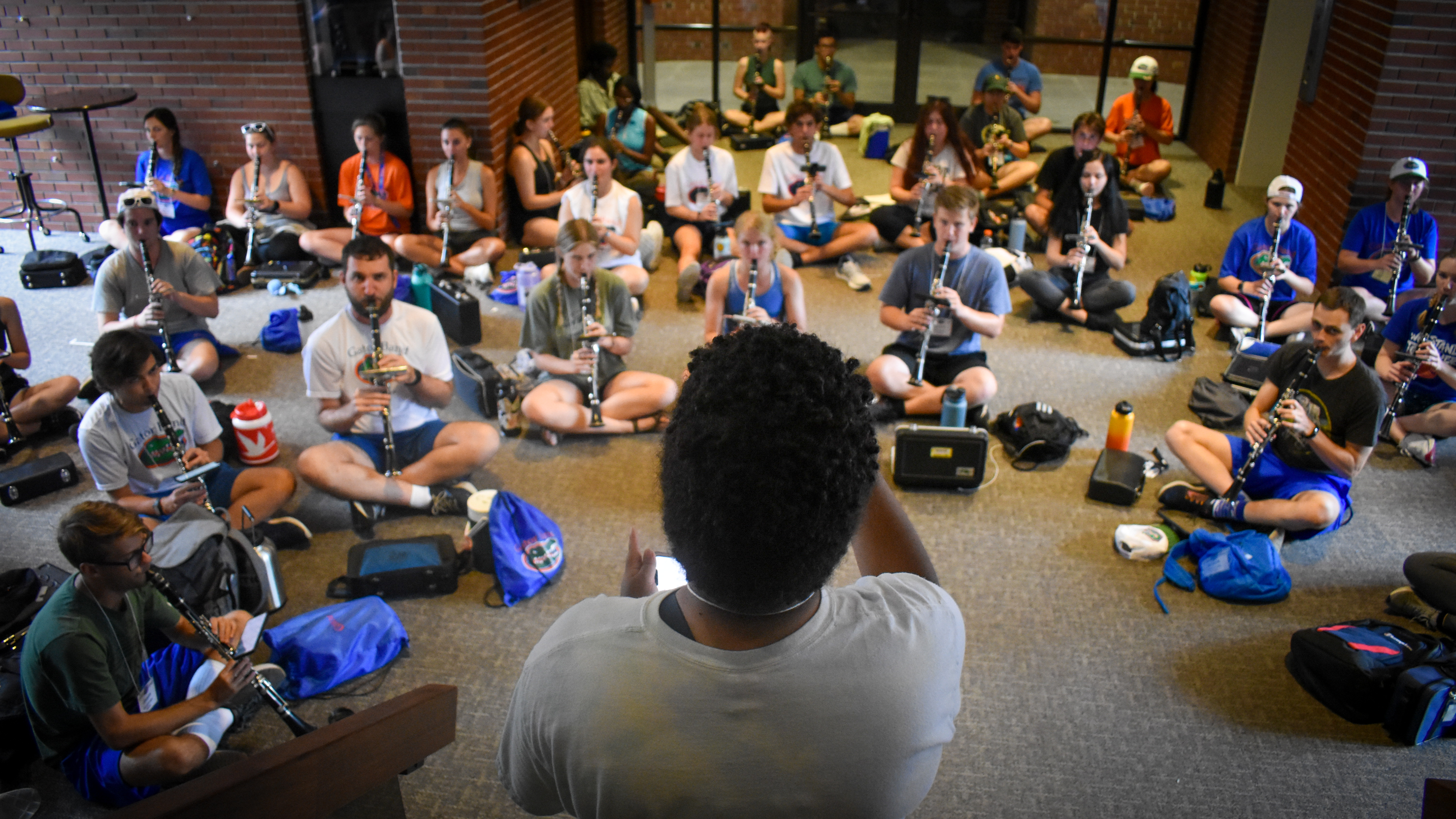 An conductor in the foreground waves her arms at a group of sitting musicians.