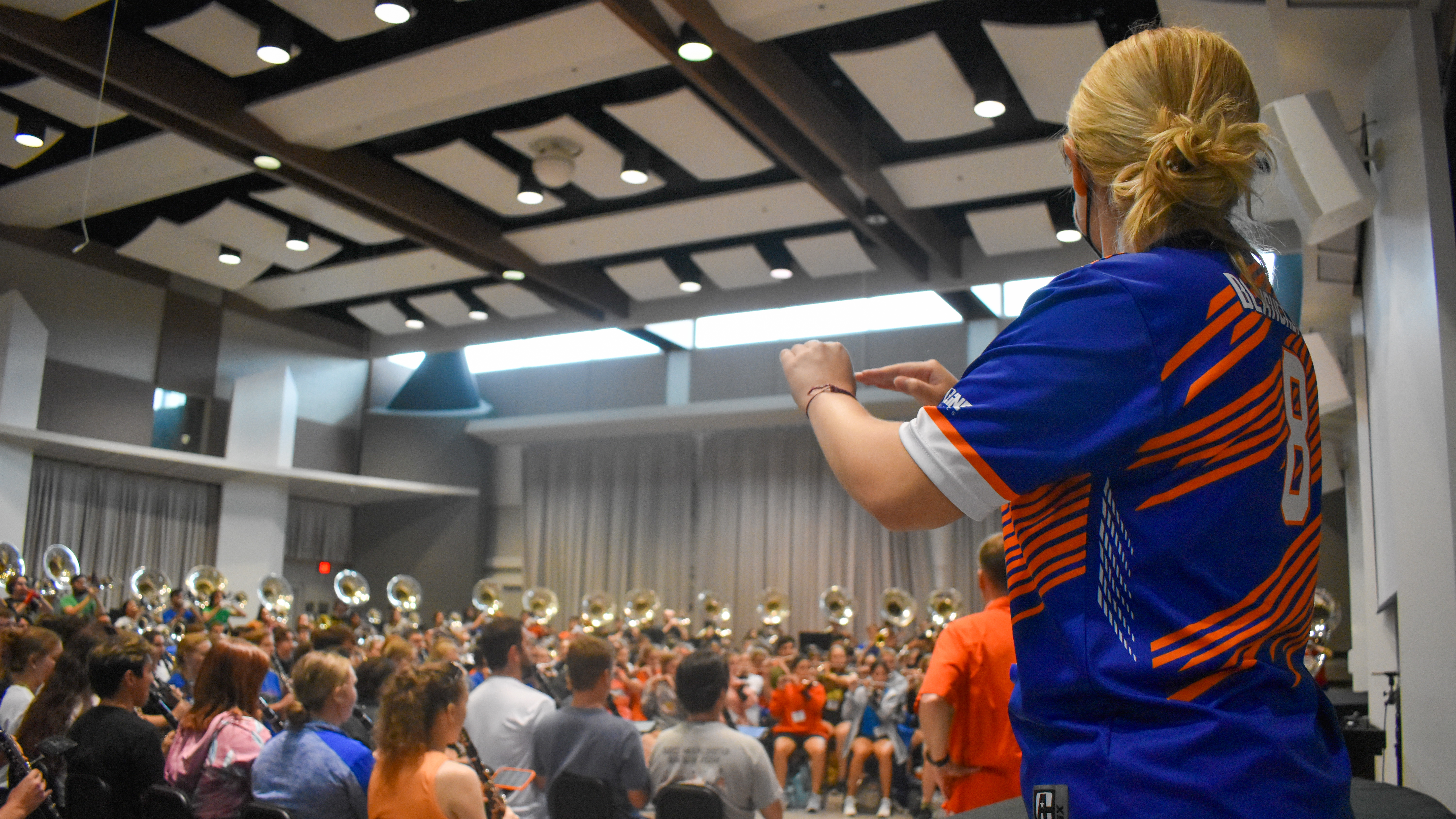 A drum major conducts a band sitting in a band hall.