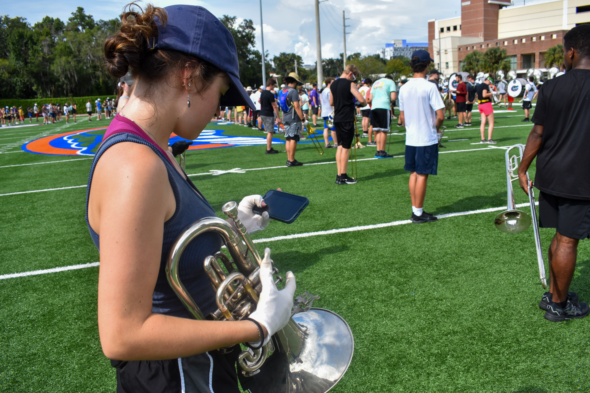 A student holding a mellophone looks at her phone on a football field.