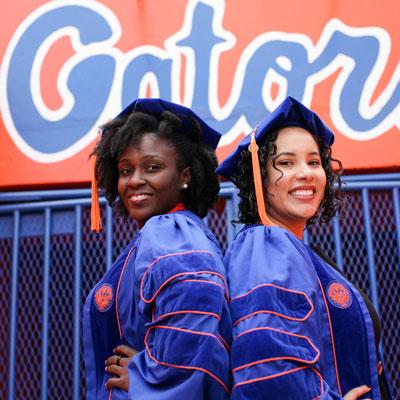 Two woman in graduation regalia pose together. 