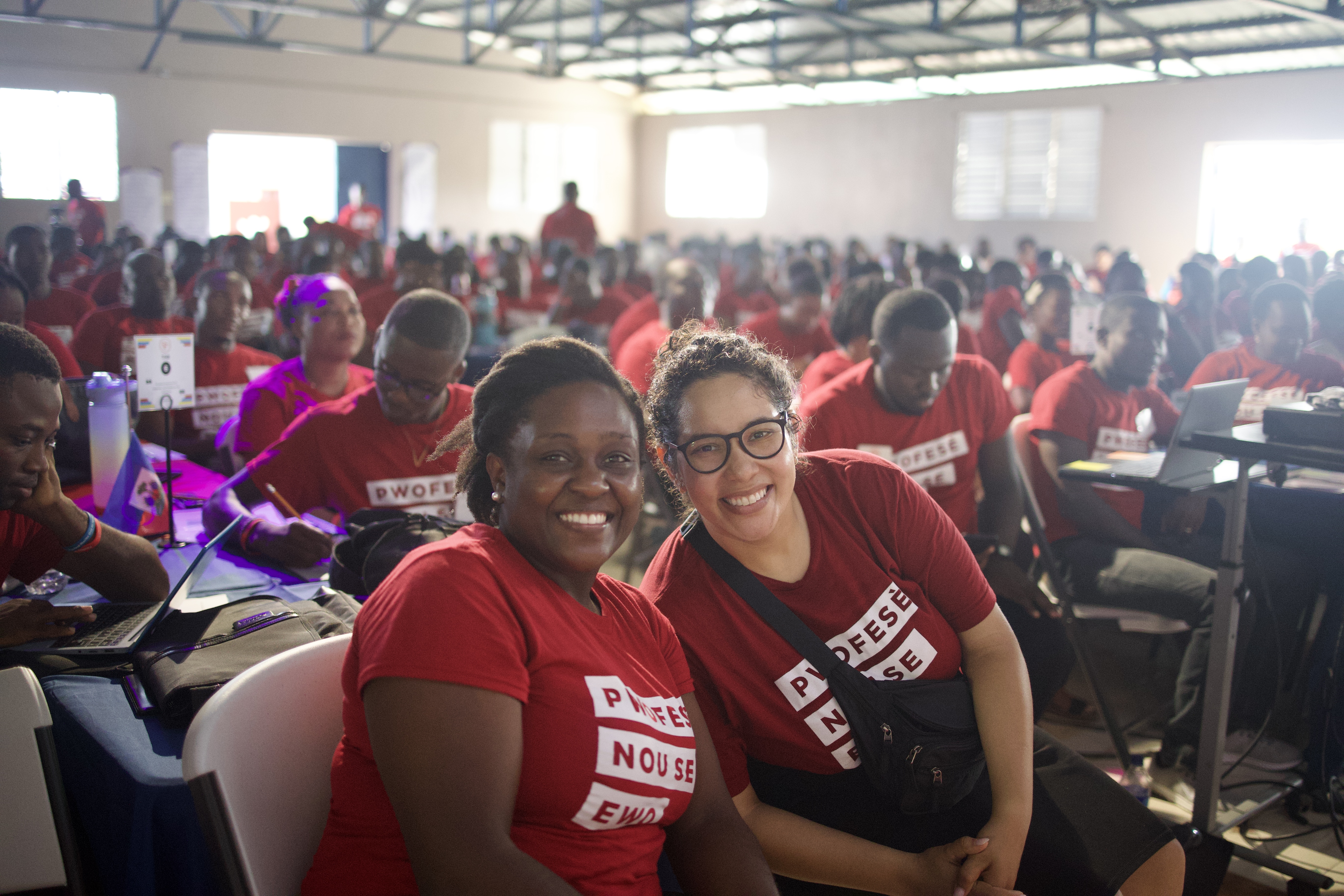 Two women in red shirts pose in a room full of people. 