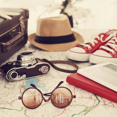 A stock image of travel accessories including a brown suitcase, tan fedora hat, red sneakers, camera, sunglasses, book and tablet sitting on top of a map.