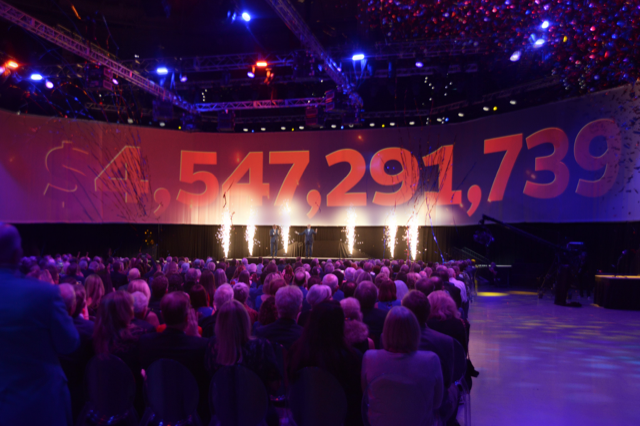 A crowd looking at a stage with fire on it, orange and blue lights, and a large number projected on a screen. 