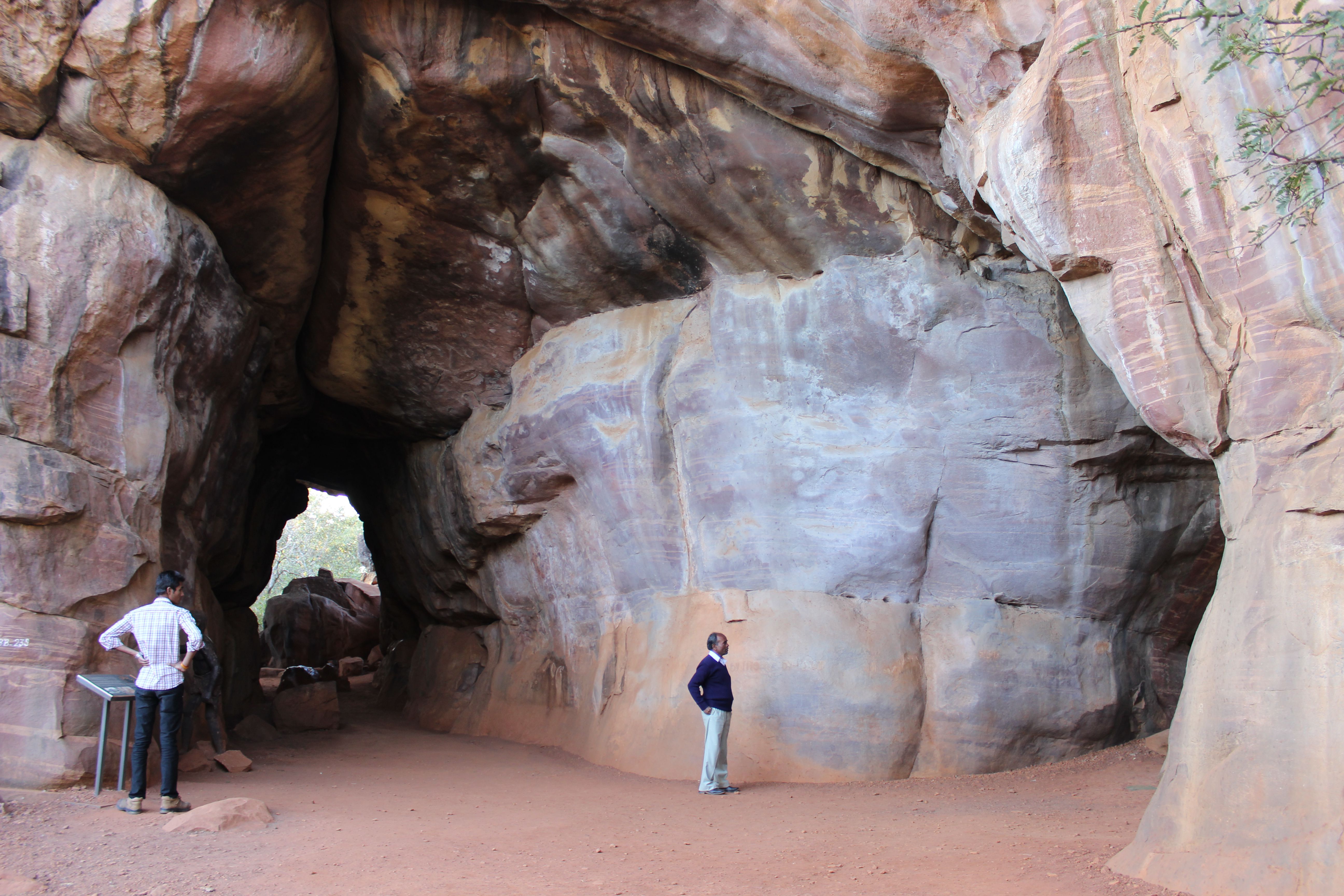 Two people stand near entrances to rock caves that loom over them