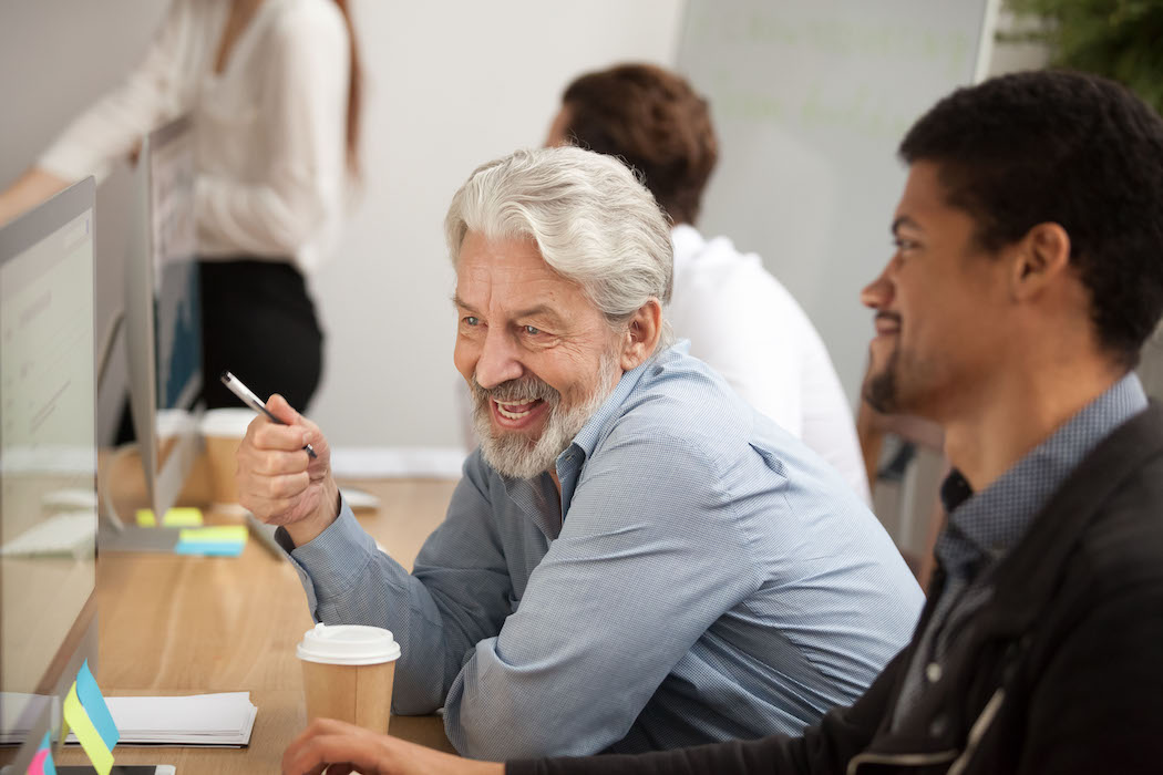 An older man working in an office gestures toward a work project as his younger colleague looks on