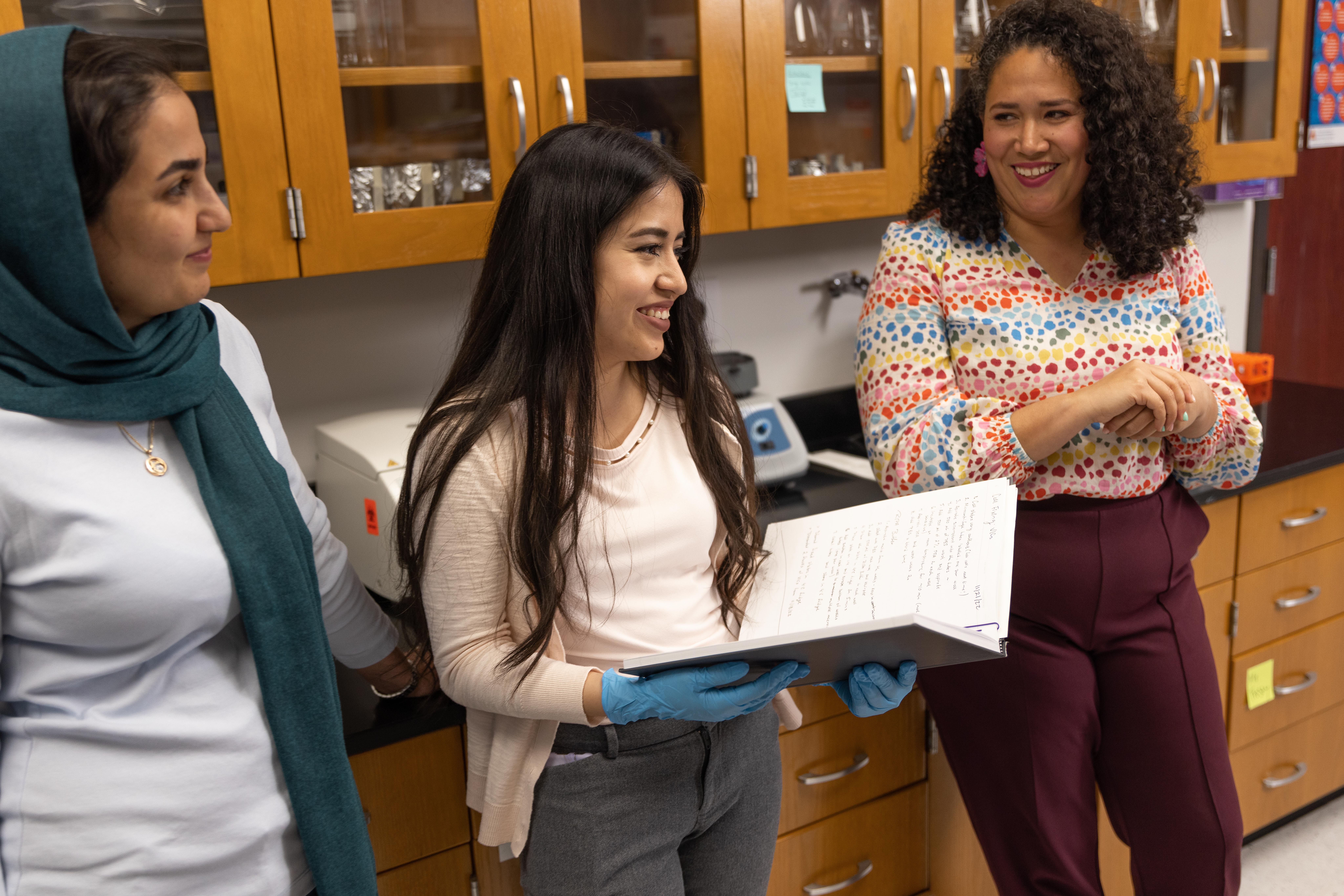 Three females are standing in a lab. The first female is wearing a hijab, the second female is holding a book and the third is smiling and glancing at the others.