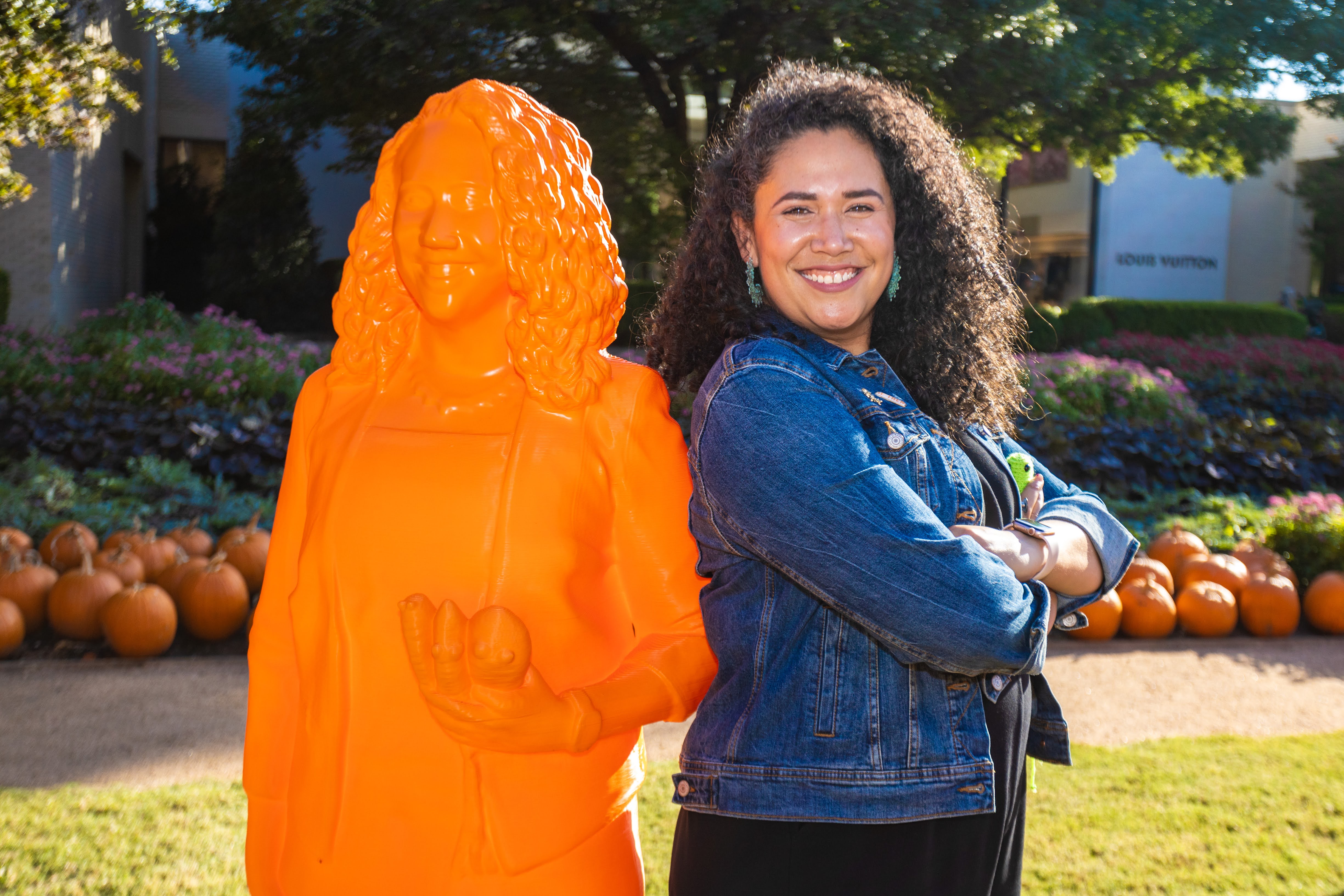 A female standing with crossed arms next to an orange statue of herself. 