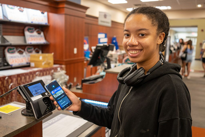 A young woman holds a phone near a contactless payment reader at the UF Bookstore.
