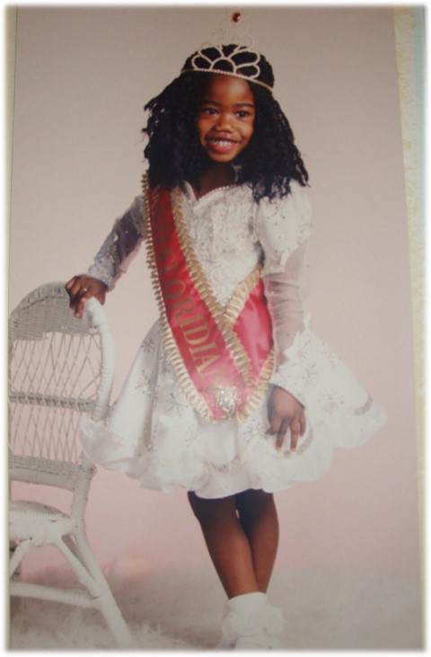 A girl in a pageant dress, wearing a crown and a sash, leans on a chair.