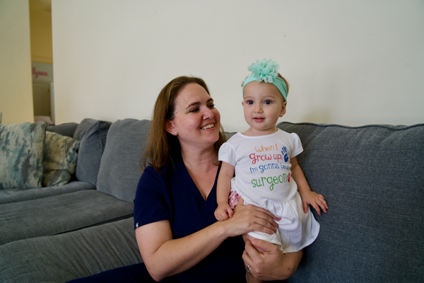 A doctor in scrubs holds her child on a couch.