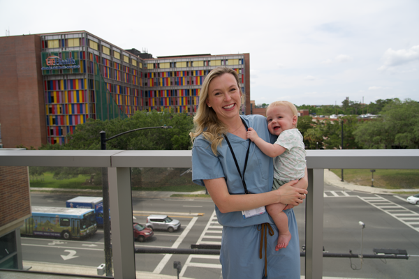 A doctor in scrubs holds her child with UF Health Shands in the background.