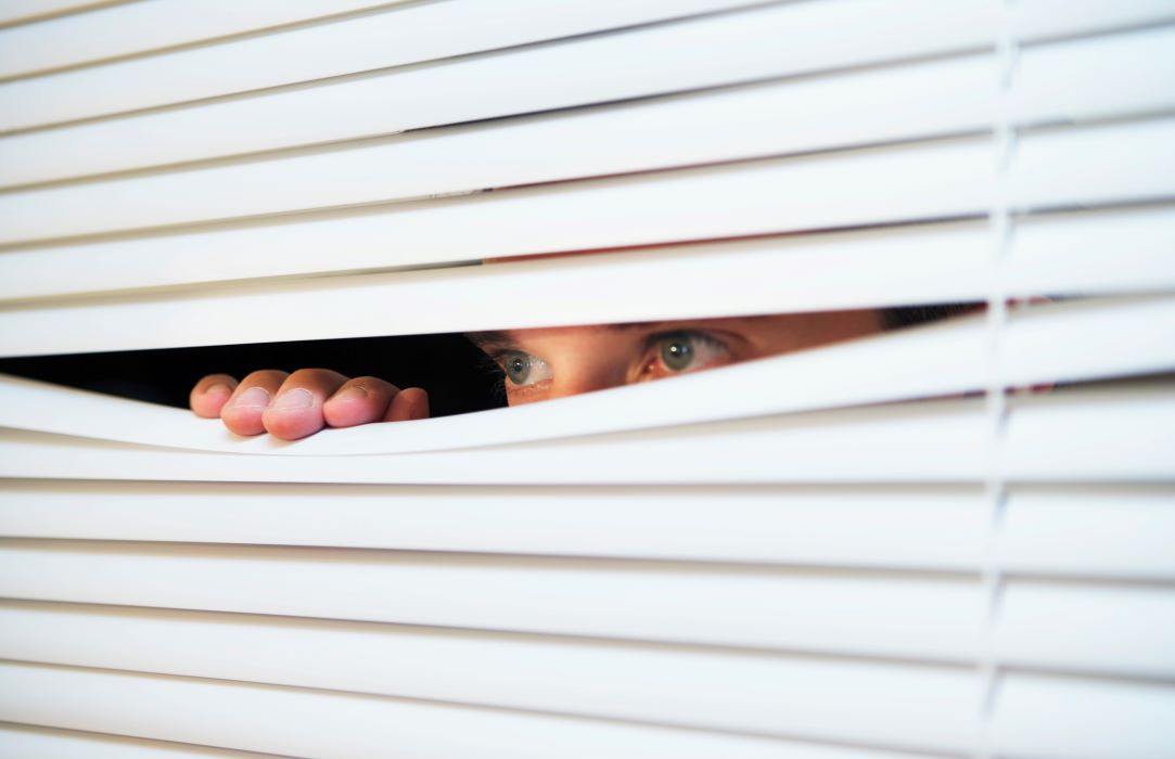 A man's eyes peek from behind venetian blinds as his hand pulls the blinds apart