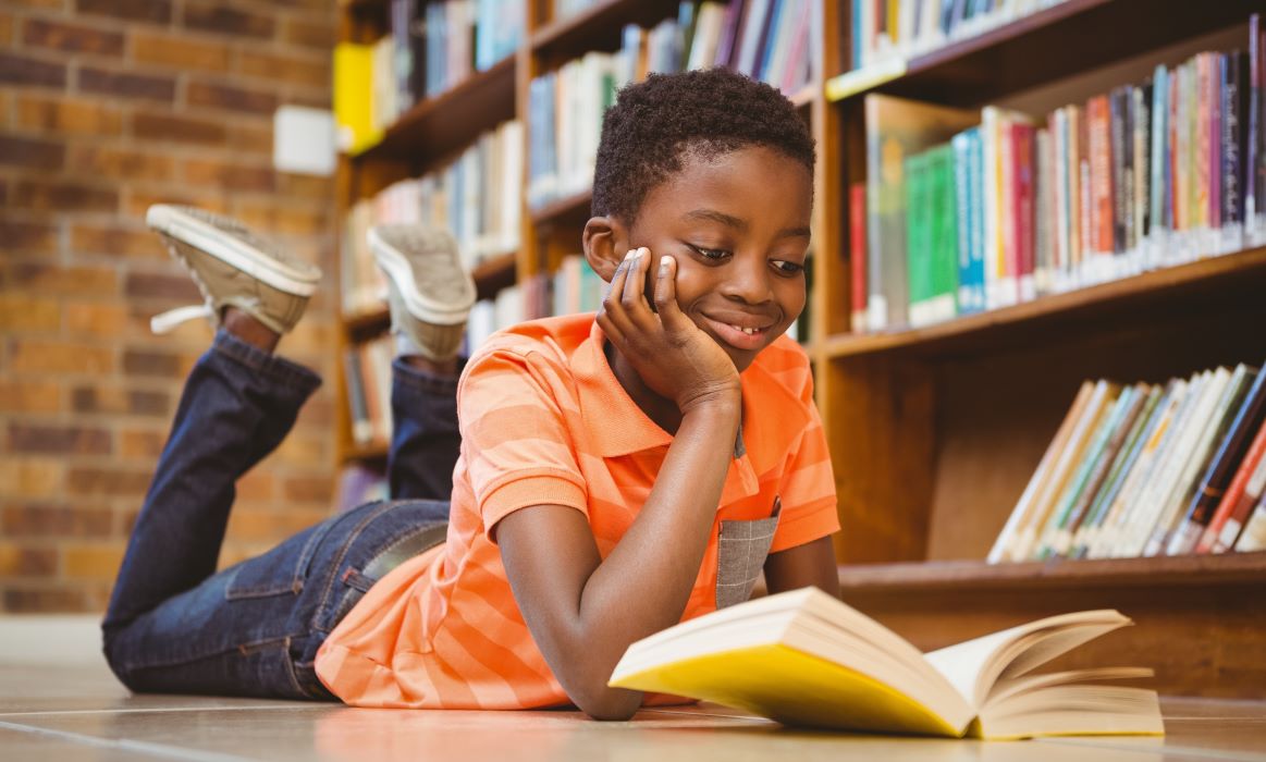 A child lays on the ground of a library smiling while reading an open book
