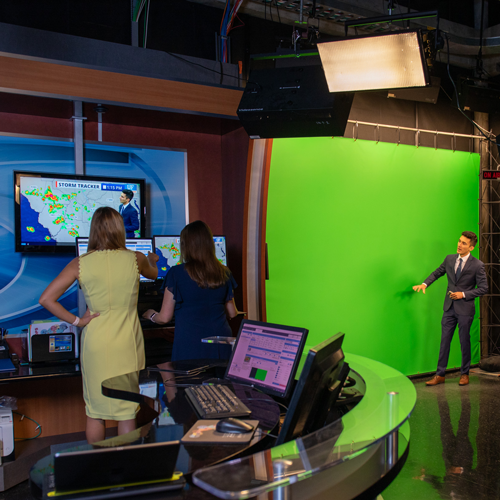 A meteorologist stands in front of a green screen pointing at things while two people watch his broadcast.