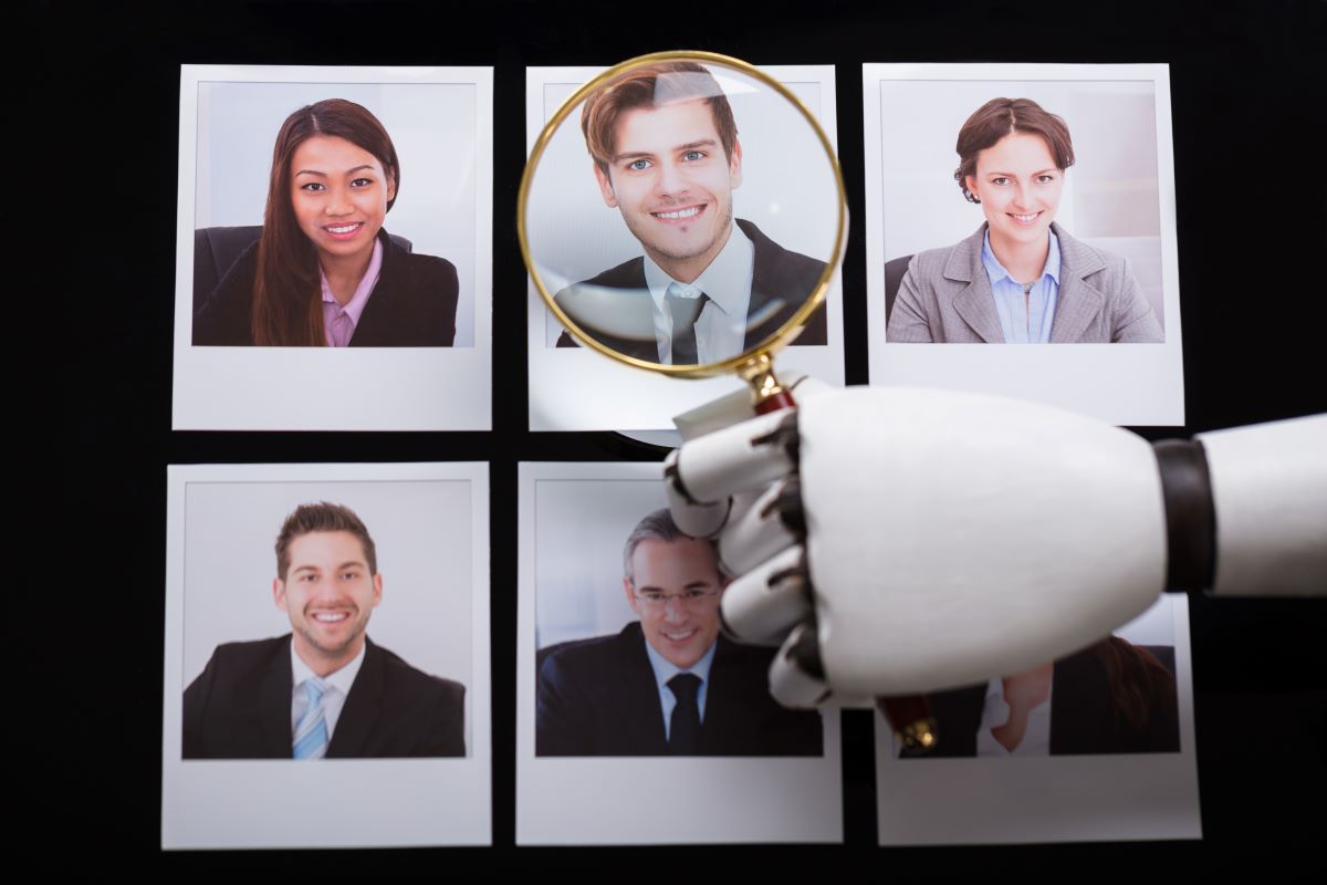 A robotic hand holds a magnifying glass over a set of six polaroid pictures of job candidates