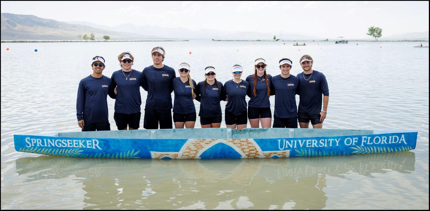 A team of college students facing the camera with their arms behind each others backs behind a blue-painted concrete canoe floating in the water