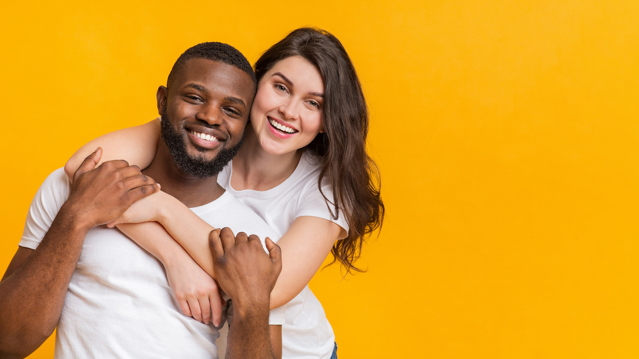 An adult couple facing the camera with the woman's arms around the man's chest set against a pure yellow background