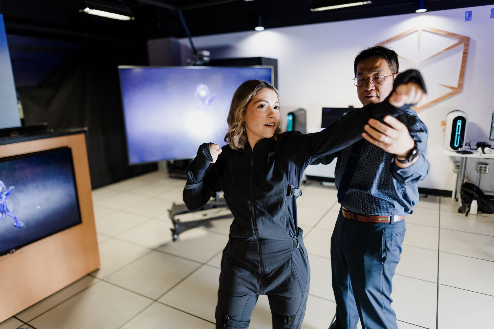 A woman in a jumpsuit punches the air guided by a man standing next to her with computer screens behind them.