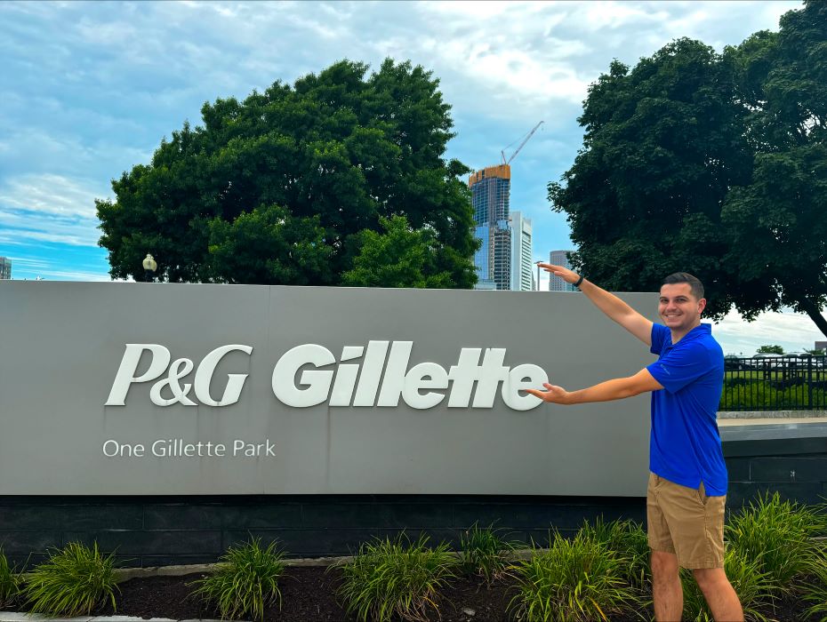 A student in a blue polo makes the Gator chomp hand motion in front of a sign that says P&G Gillette