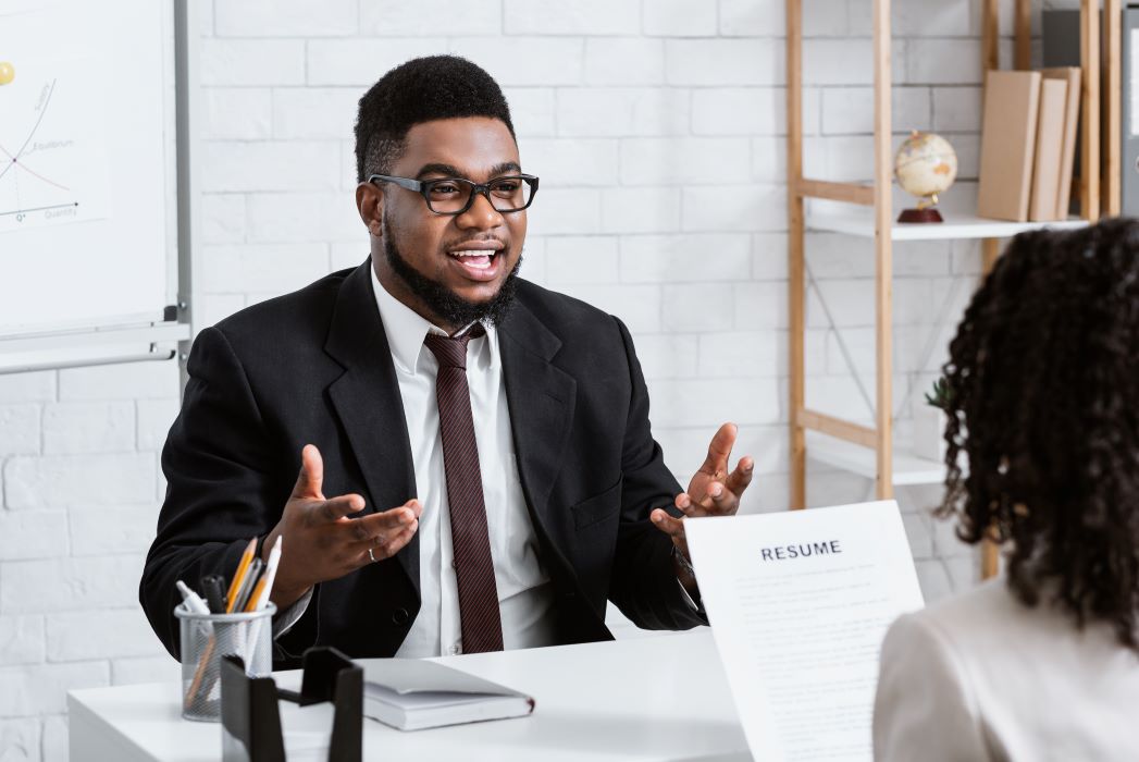 A job candidate sits at a desk and gestures toward a hiring manager