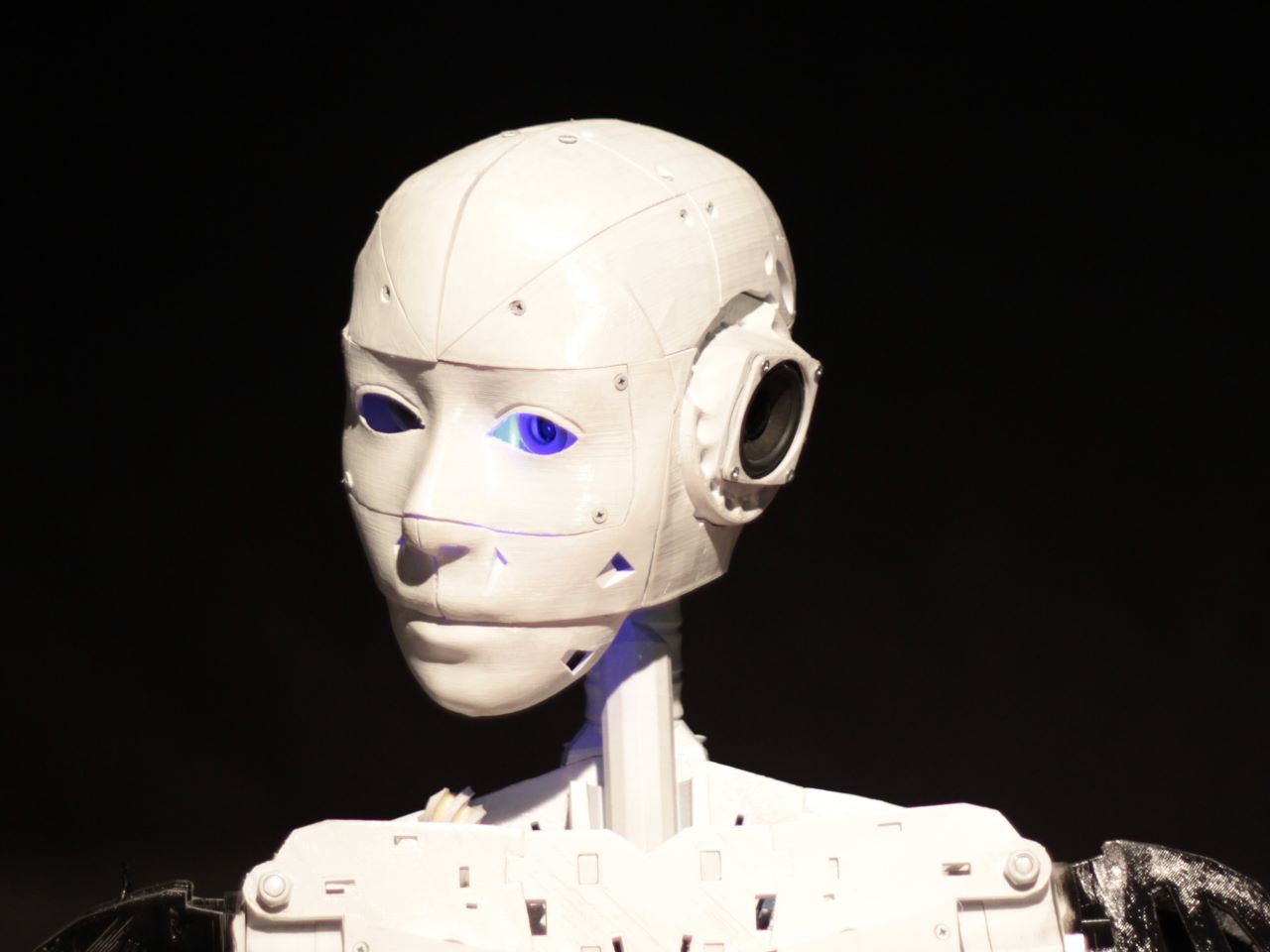 The head of a robot with metal casing but a lifelike face against a black background