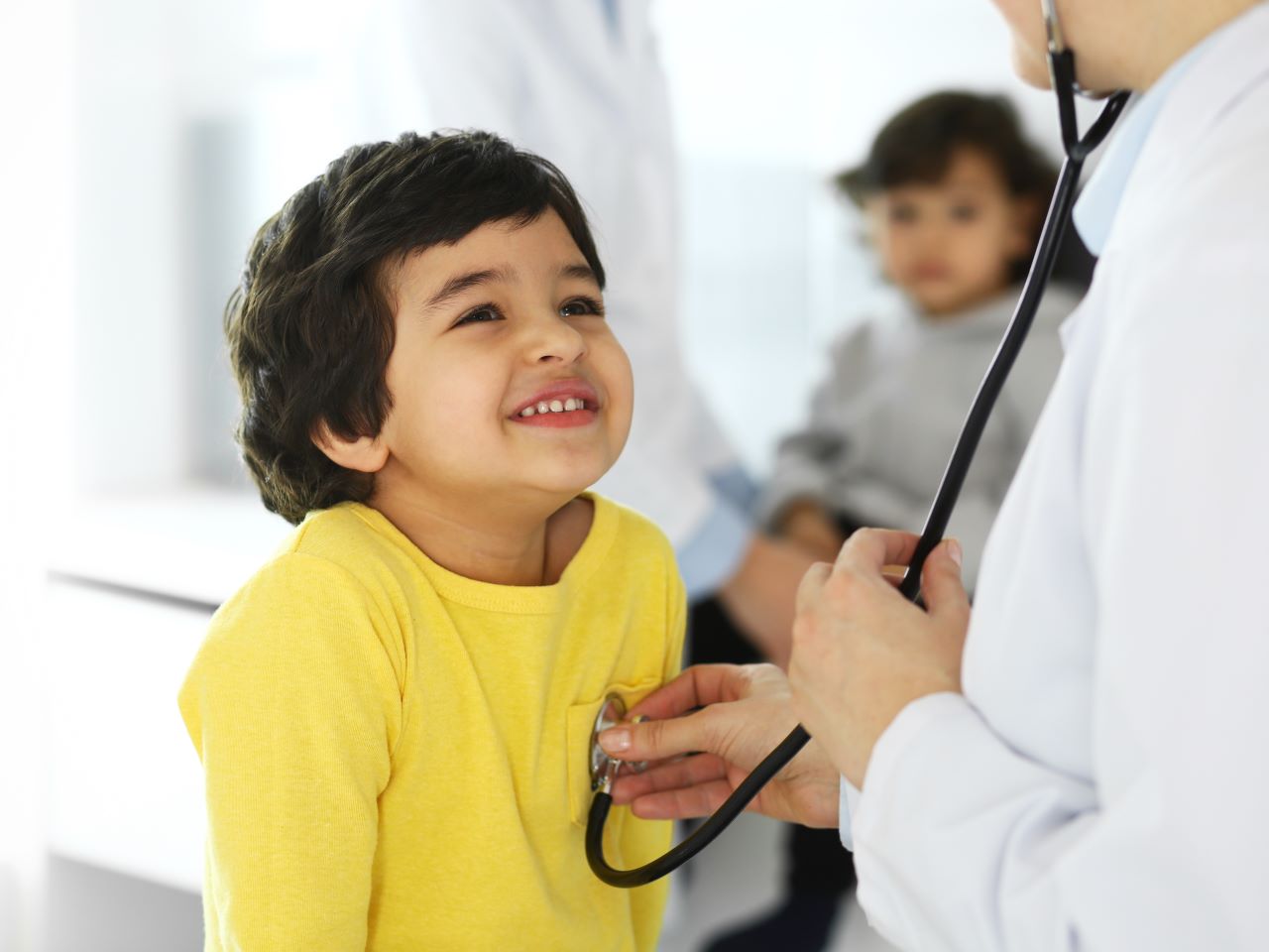 A young child smiles up at a doctor who examines his chest with a stethoscope