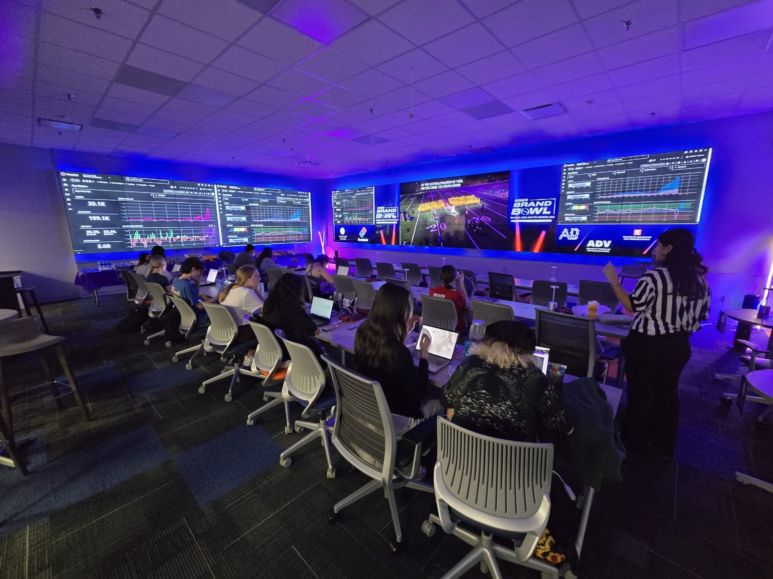 Students sit at computers in a dim classroom surrounded by televisions showing the Super Bowl.