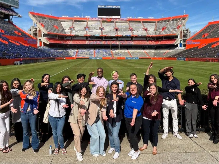 A dozen students make the Gator chomp hand sign on the field of the University of Florida football stadium